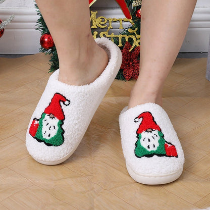 Chaussons de Noël Doudounea blancs douillets avec motif lutin festif, photographiés devant un décor de Noël avec des ornements rouges et verts